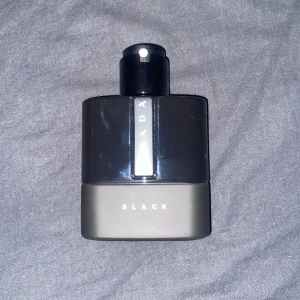 Prada Black parfym - Säljer en elegant och stilren Prada Black parfym. Flaskan har en modern design med en svart överdel och en matt grå bas. Perfekt för den som söker en sofistikerad doft med en touch av mystik.
