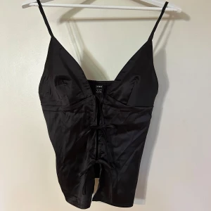 Svart satinlinnetopp från Shein - Snygg svart satinlinnetopp från Shein med smala axelband och knytning framtill. Perfekt för en stilren look med en touch av elegans. Toppen har en djup v-ringning och en figurnära passform.