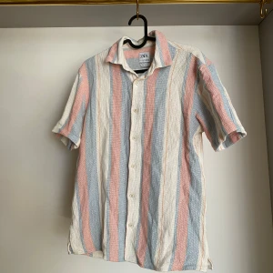 Randig kortärmad skjorta från Zara - Säljer en avslappnad randig kortärmad skjorta från Zara i mjuka pastellfärger. Skjortan har korta ärmar och knappar framtill. Perfekt för en somrig look!