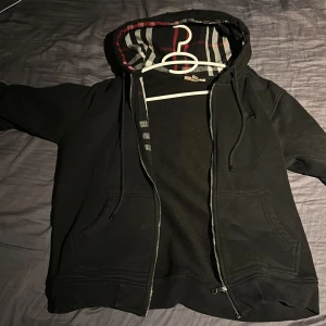 Svart huvtröja med dragkedja - Snygg svart huvtröja från Burberry med dragkedja och randigt mönster i luvan. Perfekt för en casual look med stil. Den har långa ärmar och en broderad logga på bröstet.