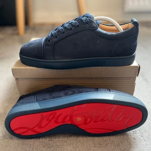 Mörkblå sneakers från Christian Louboutin - Snygga mörkblå sneakers från Christian Louboutin med den ikoniska röda sulan. Skorna har snörning och är tillverkade i mocka, vilket ger en lyxig känsla. Perfekta för dig som vill ha en stilren look med en touch av exklusivitet. obs! sko paketet är skadat