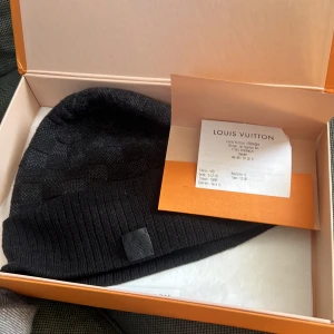 Svart mössa från Louis Vuitton - Snygg svart mössa från Louis Vuitton med ribbad kant. Tillverkad i Italien, perfekt för att hålla dig varm och stilren under kyliga dagar. Diskret logotypdetalj framtill. Box kvitto allting finns med