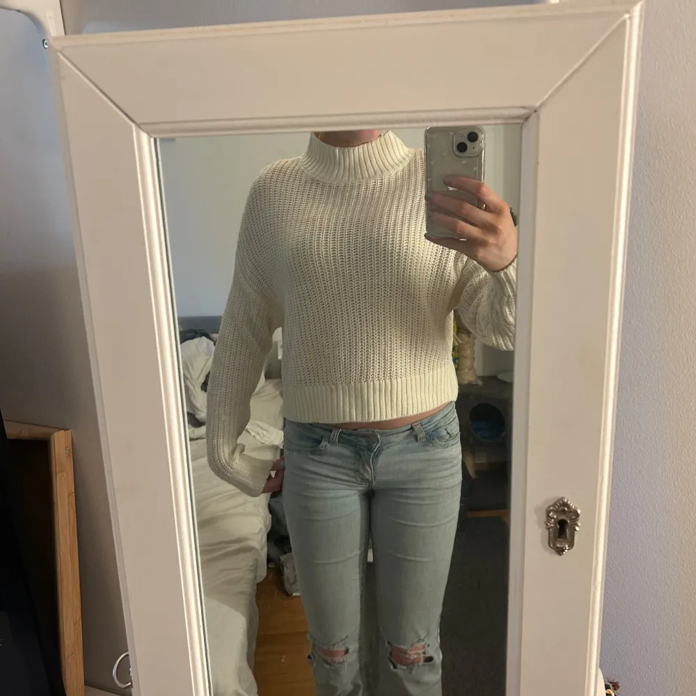 Säljer en stilren vit stickad tröja med hög krage. Tröjan är croppad och har långa ärmar, perfekt för en trendig look. Passar bra till jeans eller kjol för en avslappnad stil.. Neuletakit & Villapaidat.