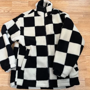 Svartvit fleecejacka från H&M - Säljer en cool svartvit fleecejacka från H&M med ett schackrutigt mönster. Jackan har dragkedja och långa ärmar, perfekt för kyliga dagar. Den är både stilren och bekväm. Säljer då den ej kommit till användning 