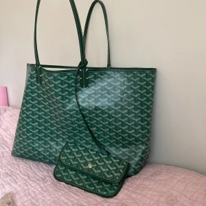 Grön mönstrad väska  - Snygg grön shoppingväska med ikoniskt mönster. Väskan har långa handtag och en matchande liten plånbok. Perfekt för att bära med sig allt man behöver under dagen. Den har en öppen design med rymligt inre.