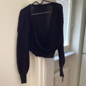 Svart stickad omlottkofta - Säljer en svart stickad omlottkofta med långa ärmar. Perfekt för kyligare dagar och ger en stilren look. Koftan har en djup omlottdesign framtill.