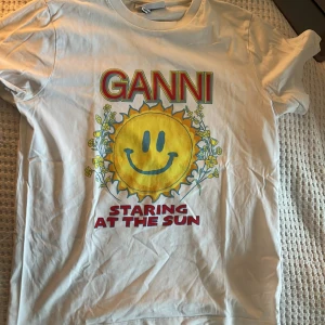 Vit t-shirt med soltryck från Ganni - Säljer en vit t-shirt från Ganni med ett färgglatt tryck av en sol med ett leende ansikte och texten 'Staring at the Sun'. Perfekt för en solig dag eller för att lysa upp vardagen. T-shirten har korta ärmar och är gjord i mjuk bomull.