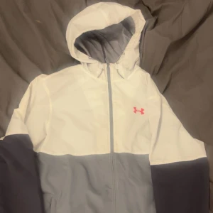 Vit och grå vindjacka från Under Armour - Säljer en snygg vit och grå vindjacka från Under Armour. Jackan har en dragkedja framtill och en justerbar huva. Perfekt för blåsiga dagar och har en sportig look med det röda logotypmärket på bröstet.