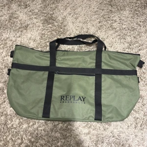 Replay Travel Bag - Säljer en grön väska från Replay med svarta detaljer och dragkedja. Perfekt för att bära med sig allt du behöver. Väskan har ett stort huvudfack och en mindre ficka med dragkedja på insidan. Aldrig använt