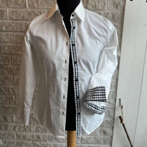 Vit skjorta från Ernst Alexis - Snygg vit skjorta från Ernst Alexis med svartvitrutig detalj längs knäppningen och på insidan av manschetterna. Skjortan har långa ärmar och knappar framtill. Perfekt för en stilren look.