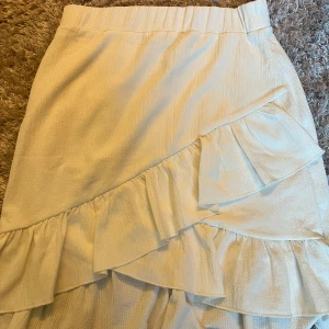 Vit kjol med volangdetaljer - Säljer en stilren vit kjol med volangdetaljer. Kjolen har en omlott-design och är perfekt för en elegant look. Den är gjord i ett lätt och bekvämt material.