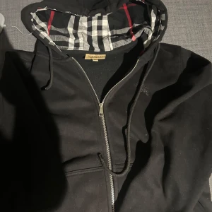 Svart zip från Burberry - Säljer en stilren svart hoodie från Burberry med dragkedja och klassiskt rutigt mönster i huvan. Perfekt för en avslappnad look med en touch av lyx. Huvan har snörning och jackan har långa ärmar.