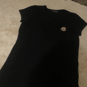 Svart t-shirt från Moncler - Säljer en stilren svart t-shirt från Moncler med korta ärmar och en liten broderad logga på bröstet. Perfekt för en casual look eller att matcha med dina favoritjeans.