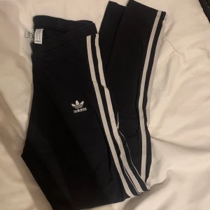 Svarta byxor tights från Adidas - Säljer ett par svarta tights från Adidas med tre vita ränderna längs sidorna. Byxorna har en tight passform och är perfekta även för träning. 