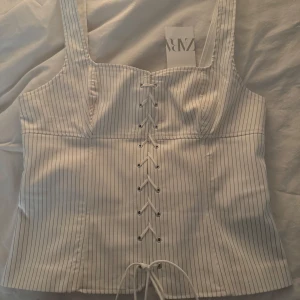Vit randig korsett från Zara - Snygg vit korsett från Zara med smala svarta ränder och snörning framtill. Den har breda axelband och en dragkedja bak för enkel påklädning. Perfekt för en stilren look. Aldrig använd💕Storlek XL men passar L också