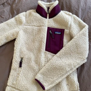 Vit teddyjacka från Patagonia - Säljer en supermysig vit teddyjacka från Patagonia med lila detaljer. Jackan har en dragkedja framtill och en praktisk bröstficka med dragkedja. Perfekt för kyliga dagar! 🐻