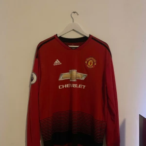 Röd Manchester United fotbollströja från Adidas authentic - Säljer en röd Manchester United fotbollströja från Adidas med långa ärmar. Tröjan har klubbens emblem och sponsorlogga på framsidan samt spelarens namn och nummer på baksidan. Perfekt för fans som vill visa sitt stöd.