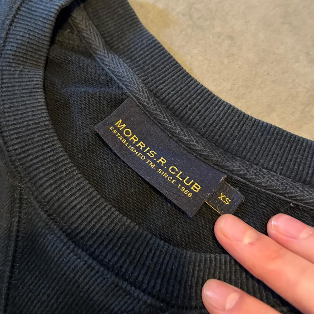 Säljer en stilren svart tröja från Morris R Club med ett broderat emblem på bröstet. Tröjan har långa ärmar och ribbade muddar vid ärmslut och nederkant. Perfekt för en avslappnad look.. Neuletakit & Villapaidat.