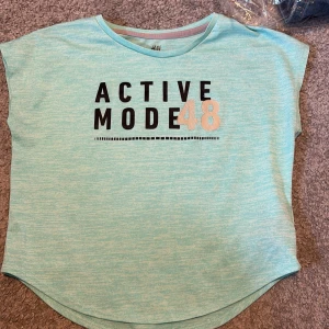 Turkos idrottströja från H&M - Säljer en turkos t-shirt från H&M med texten 'Active Mode 48' i svart och vitt på framsidan. Den har en rund halsringning och korta ärmar. Är i storlek 122-128. 