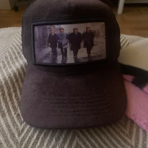 Brun keps från Maggiore - Snygg brun keps från Maggiore med ett unikt tryck av fyra personer på framsidan. Kepsen har en justerbar rem baktill för perfekt passform. Perfekt för en cool och avslappnad stil. Inte använd en endaste gång. 