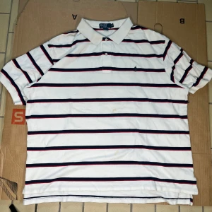 Vit randig pikétröja från Ralph Lauren - Säljer en klassisk vit pikétröja från Ralph Lauren med marinblå och röda ränder. Tröjan har korta ärmar och en krage med knappar. Perfekt för en stilren look. Tillverkad i 100% bomull. Har ni några frågar fråga gärna oss! Priset kan alltid förhandlas 