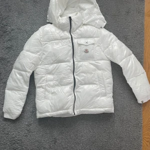 Vit dunjacka från Moncler - Säljer en snygg vit dunjacka från Moncler med huva och dragkedja. Jackan har en bröstficka med knapp och Moncler-logga. Perfekt för kyliga dagar!