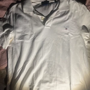 Vit pikétröja från GANT - Säljer en klassisk vit pikétröja från GANT i storlek M. Tröjan har korta ärmar och en broderad logga på bröstet. Perfekt för en stilren och avslappnad look. Hör av dig vid intresse!