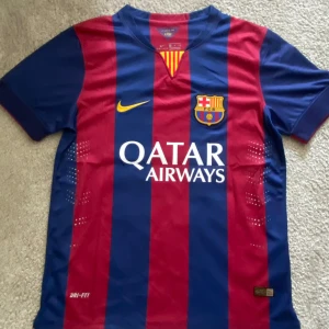 Barcelona 14/15, Messi 10  - Säljer en officiell FC Barcelona fotbollströja från Nike med Messi och nummer 10 på ryggen. Tröjan är randig i blått och rött med Qatar Airways och UNICEF-loggor. Den har korta ärmar och är perfekt för alla Barça-fans!