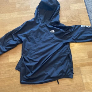 Svart vindjacka från The North Face - Säljer en svart vindjacka från The North Face med huva och dragkedja. Jackan har logotypen tryckt på bröstet och ärmen. Perfekt för blåsiga dagar och lätt att packa med sig. Pris kan diskuteras och byten.