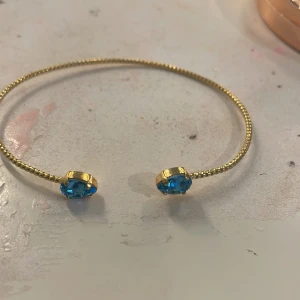 Guldarmband med blå stenar - Elegant guldarmband med två gnistrande blå stenar i ändarna. Armbandet har en tunn och flexibel design som gör det enkelt att bära. Perfekt för att lägga till en färgklick till din outfit.