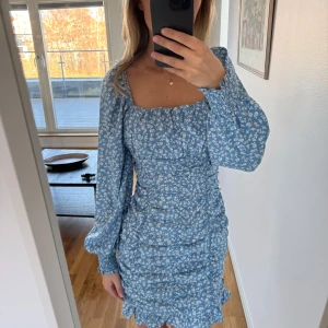Blå blommig klänning - Säljer en söt blå klänning med vita blommor. Klänningen har långa ärmar och en volangdetalj vid fållen. Perfekt för vår och sommar! Endast använd 1 gång!