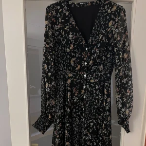 Blommig långklänning från Gina Tricot - Snygg långklänning från Gina Tricot med ett blommigt mönster på svart bakgrund. Klänningen har långa ärmar och knappar framtill, vilket ger en elegant touch. Perfekt för både vardag och speciella tillfällen.