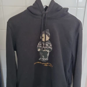 Svart hoodie med Polo Bear från Ralph Lauren - Säljer en svart hoodie från Ralph Lauren med det ikoniska Polo Bear-motivet på framsidan. Tröjan har en klassisk huva med dragsko och är perfekt för en avslappnad stil. Den är både stilren och bekväm.