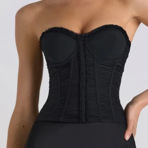 Svart korsett med snörning - Snygg svart korsett med snörning i ryggen och dekorativa detaljer framtill. Axelbandslös design som ger en elegant look. Perfekt för att skapa en stilren och sofistikerad outfit.
