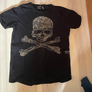 Svart t-shirt med dödskalle från Rusty Neal - Säljer en svart t-shirt från Rusty Neal med ett stort dödskalletryck på framsidan. T-shirten har en normal passform och korta ärmar. Perfekt för en cool och avslappnad stil. 🖤