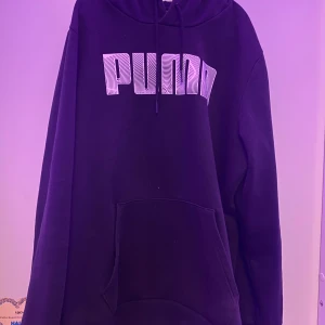 Svart hoodie från Puma - Säljer en svart hoodie från Puma med stor logga på bröstet. Den har en klassisk känguruficka och justerbar huva med snören. Perfekt för en avslappnad stil.