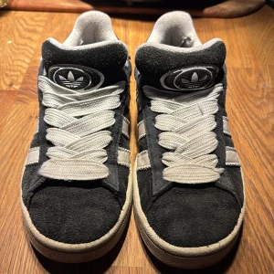 Svarta Adidas Campus sneakers  - Säljer ett par klassiska svarta Adidas Campus sneakers med vita detaljer och snörning. Skorna har en stilren design med tre vita ränder på sidan och Adidas-logga på hälen. Perfekta för en avslappnad stil. Får även med ett par svarta snören