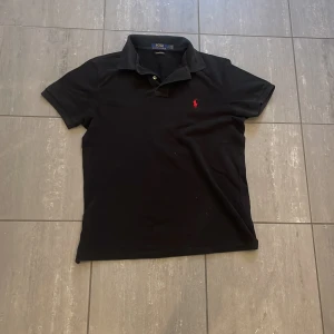 Svart pikétröja från Ralph Lauren - Säljer en stilren svart pikétröja från Ralph Lauren med röd logga på bröstet. Tröjan har korta ärmar och en klassisk krage med knappar. Perfekt för en avslappnad men ändå snygg look. Model 179cm och 65kg. Nypris 1000kr
