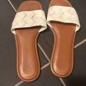 Vita sandaler från Aldo - Snygga vita sandaler från Aldo med flätad rem och öppen tå. Perfekta för varma sommardagar och ger en stilren look. Sandalerna har en brun sula som kontrasterar fint mot den vita remmen.