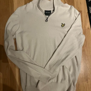Lyle scott tröja - Säljer en stilren beige tröja från Lyle & Scott med dragkedja och ribbad krage. Tröjan har ett broderat gult emblem på bröstet och långa ärmar. Perfekt för en avslappnad look. Ny skick