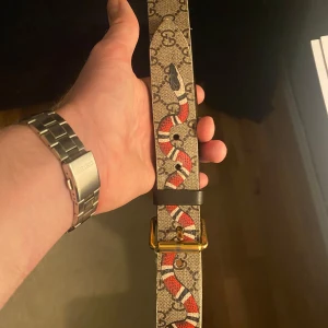 Mönstrat bälte från Gucci - Säljer ett stilrent bälte från Gucci med ett unikt mönster och en iögonfallande orm-design. Bältet har en guldfärgad spänne och är tillverkat i ett slitstarkt material med läderdetaljer. Perfekt för att ge din outfit en lyxig touch. Bältet är i storlek 85cm och är köpt för cirka 3 år sedan på Gucci i Milano.