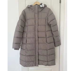jacka - Säljer en stilren beige dunjacka med huva. Jackan har långa ärmar och en quiltad design som ger extra värme. Perfekt för kyliga dagar. HELT NY