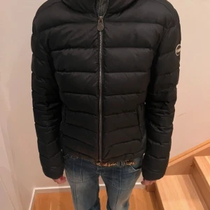 Svart dunjacka från Colmar - Snygg svart dunjacka från Colmar med dragkedja och huva. Jackan har en quiltad design och ett märke på ärmen. Perfekt för kyliga dagar.