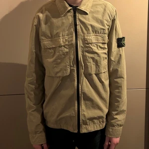 Stone Island Overshirt - Säljer en beige overshirt från Stone Island med dragkedja och två bröstfickor. Jackan har långa ärmar och en klassisk krage. Perfekt för en stilren look. Inuti finns reserv knapp ifall att någon av knapparna lossnar. Köpt på NK i Stockholm för 3 år sedan men är knappast använd utan några defekter allt, finns fler bilder om så behövs. Skicka DM för minsta lilla fundering eller för fler bilder!💯(pris kan diskuteras vid snabb affär)