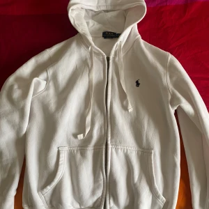 Vit hoodie från Ralph Lauren - Säljer en stilren vit hoodie från Ralph Lauren med dragkedja och justerbar huva. Den har två praktiska fickor framtill och en liten broderad logga på bröstet. Perfekt för en avslappnad look.