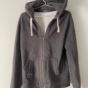 Ralph Lauren zip hoodie - Säljer en stilren grå hoodie från Ralph Lauren med dragkedja och justerbar huva. Tröjan har en liten broderad logga på bröstet och praktiska fickor framtill. Perfekt för en avslappnad look.