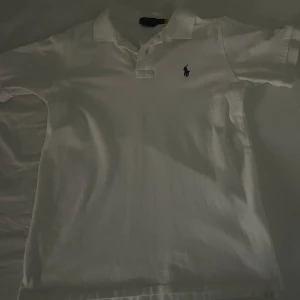 Ralph Lauren pikétröja  - Säljer en klassisk vit pikétröja från Ralph Lauren med den ikoniska loggan broderad på bröstet. Tröjan har korta ärmar och en stilren krage med knappar. Perfekt för en avslappnad men ändå stilfull look.