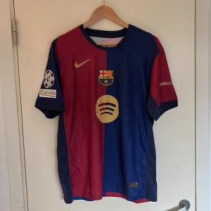 FC Barcelona fotbollströja 24/25 - Säljer en officiell FC Barcelona fotbollströja från Nike. Tröjan är i de klassiska rödblå färgerna med klubbmärket på bröstet och guldiga detaljer. Den har korta ärmar och är dekorerad med UEFA Champions League på ärmen. Perfekt för fans av laget!