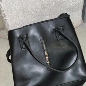 Svart axelväska med dragkedja - Snygg svart axelväska i syntetmaterial med en praktisk dragkedja framtill. Väskan har två handtag och flera fack inuti för enkel organisering. Perfekt för att bära med sig det nödvändigaste med stil.