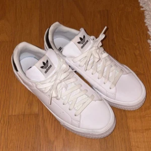 Vita sneakers från Adidas - Snygga vita sneakers från Adidas med klassisk design och svarta detaljer på hälen. Skorna har snörning och är tillverkade av syntetmaterial. Perfekta för en stilren look.